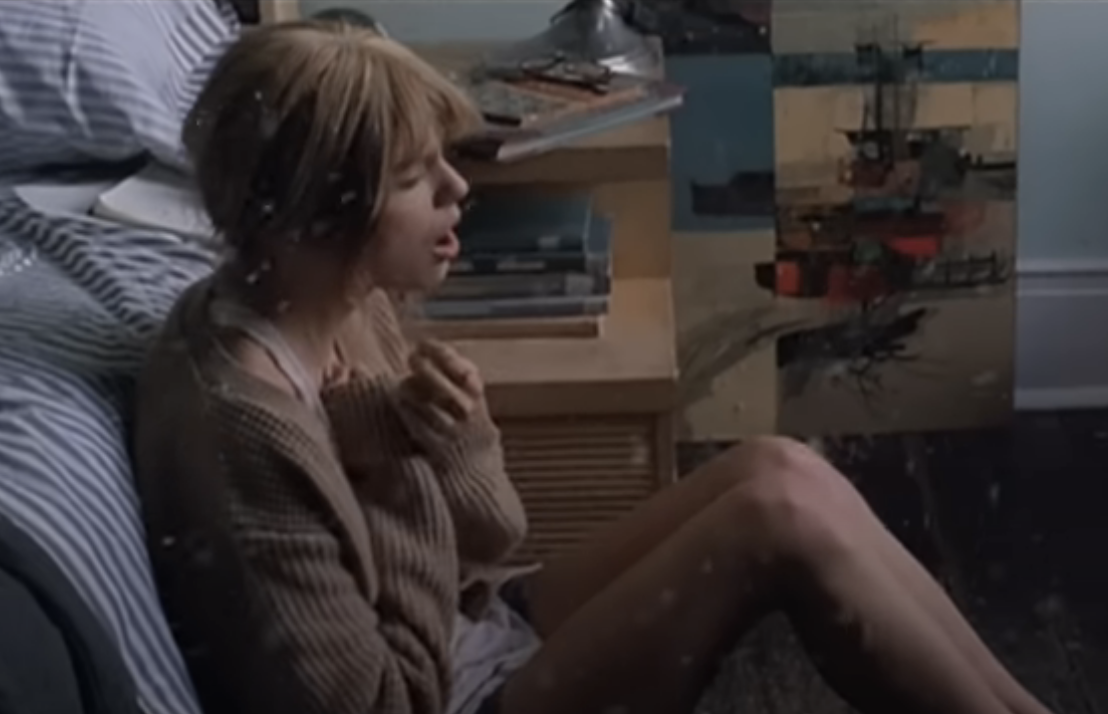 Woman in a sweater sitting on a bed with a distressed expression, clutching her chest