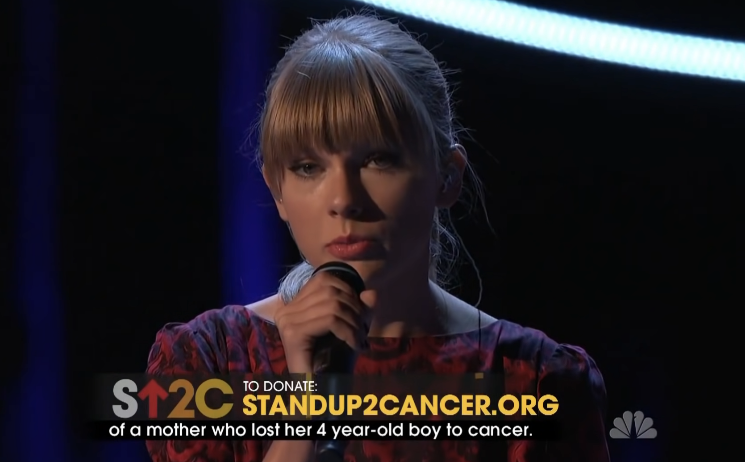Woman on stage holding microphone with &quot;StandUp2Cancer.org&quot; text and donation information on screen