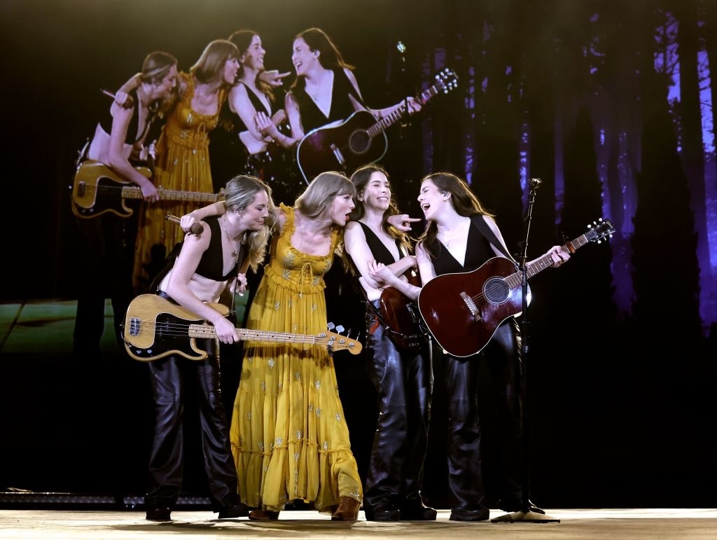 Four musicians on stage with guitars, one in a yellow dress, others in black, performing