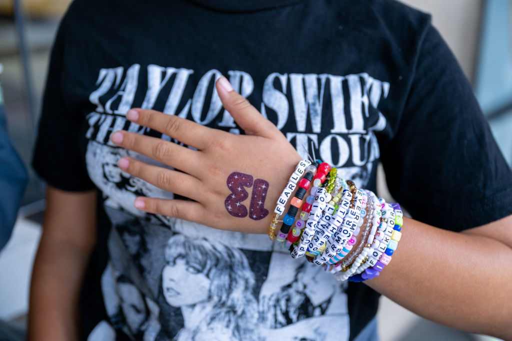 Person wearing a Taylor Swift tour shirt with hand on chest showcasing bracelets with various beads, one in the shape of the number 13