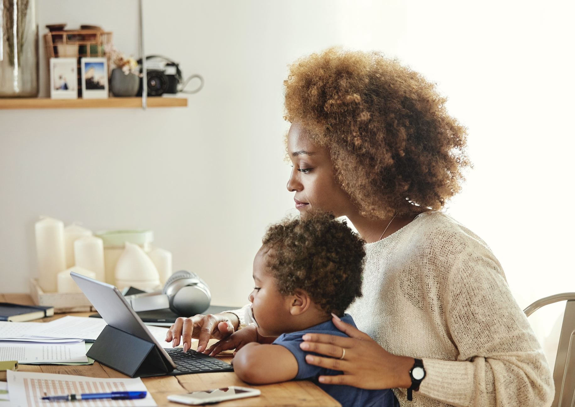 Woman works on a tablet with a young child on her lap at a home office setup