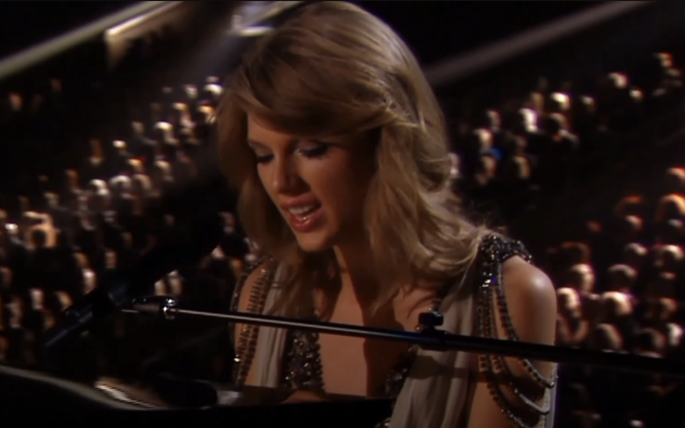 Woman with microphone playing a piano at an event, wearing a beaded dress. Audience in the background