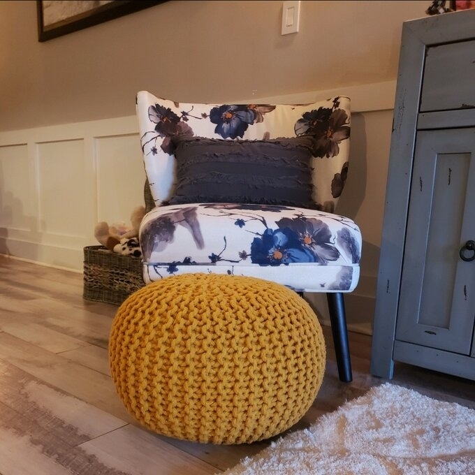 Floral-patterned armchair with a gray cushion next to a yellow round ottoman, with a wooden floor and gray cabinet
