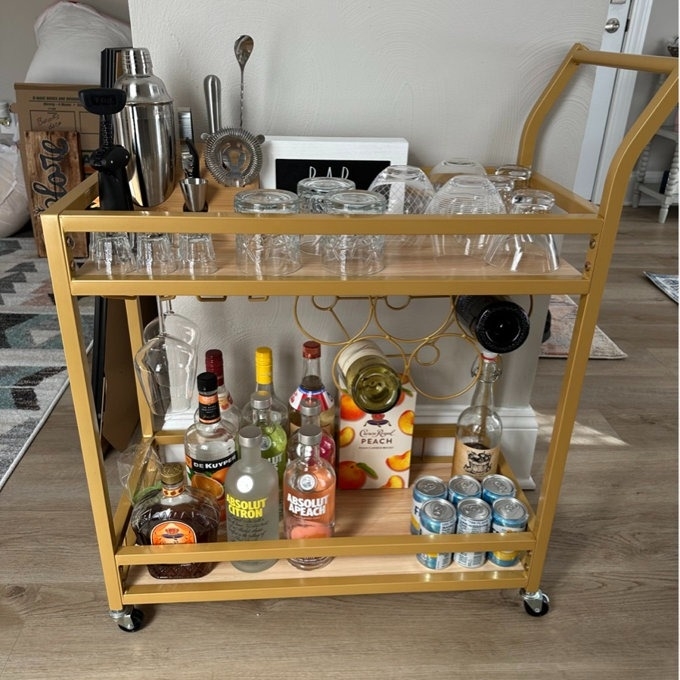 A well-stocked gold bar cart with various bottles, glasses, and canned beverages on two shelves