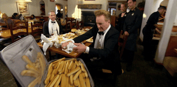 Conan O'Brien humorously throws breadsticks in a restaurant while wearing a tuxedo; staff watching