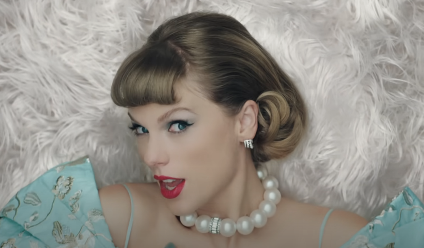 Taylor Swift lies on a white surface, wearing a vintage style dress and pearl necklace