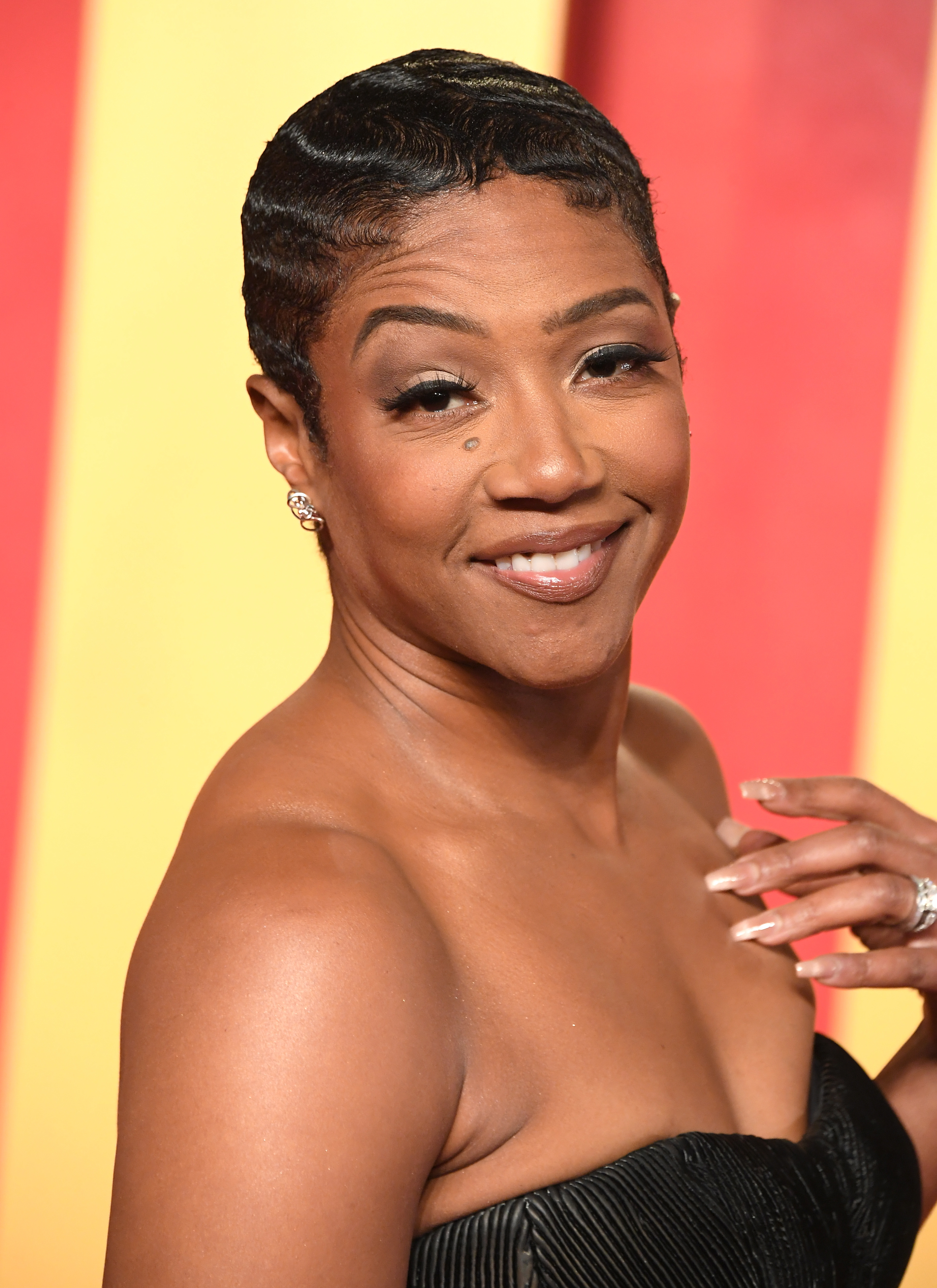 Tiffany Haddish posing with a short wavy hairstyle and a strapless ruffled dress, smiling at an event