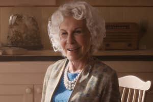 An older woman with curly hair, smiling, wearing a pearl necklace and a blue top with a patterned jacket