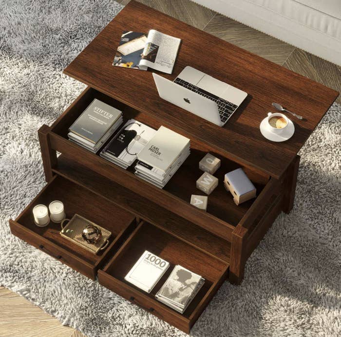 Wooden coffee table with open storage compartments, containing books and decor, with a laptop and coffee cup on top
