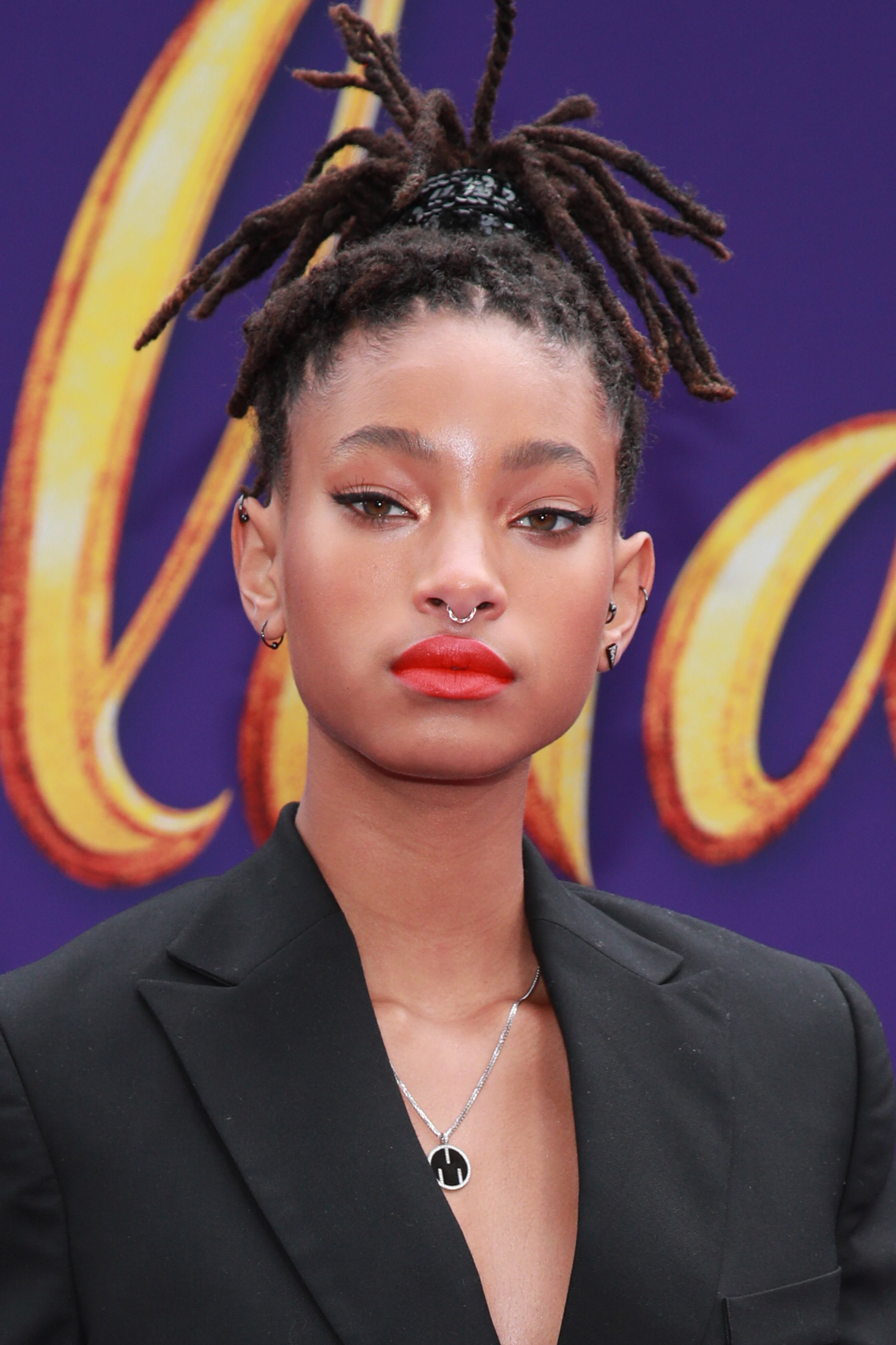 Willow Smith wearing a black blazer with a nose ring, styled hair, on a purple backdrop