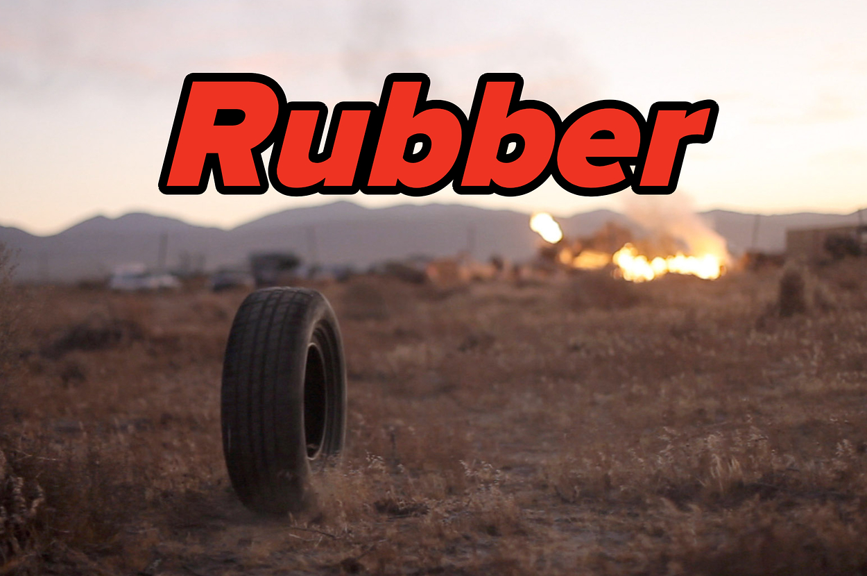 A tire stands in the foreground with "Rubber" text overlay; a fire burns in the distance