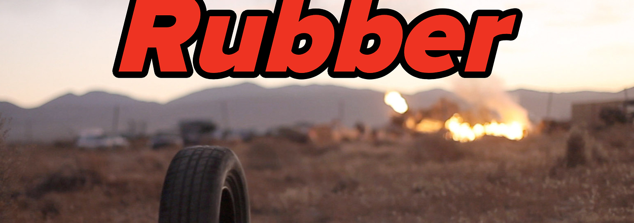 A tire stands in the foreground with "Rubber" text overlay; a fire burns in the distance