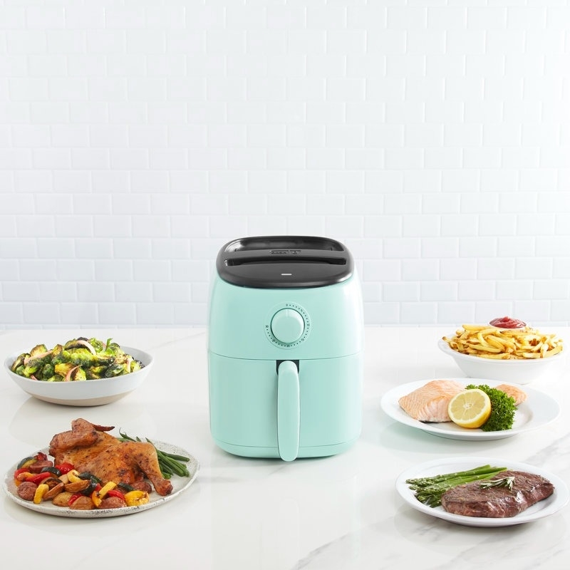 Compact air fryer on a kitchen counter surrounded by plates of cooked food