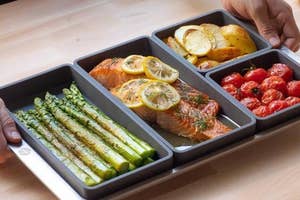 Three portioned meal prep trays with salmon, asparagus, tomatoes, and potatoes on a baking sheet