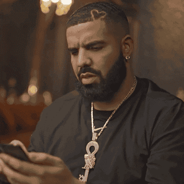 Drake looking contemplative while holding a smartphone. He&#x27;s wearing a black t-shirt and necklaces