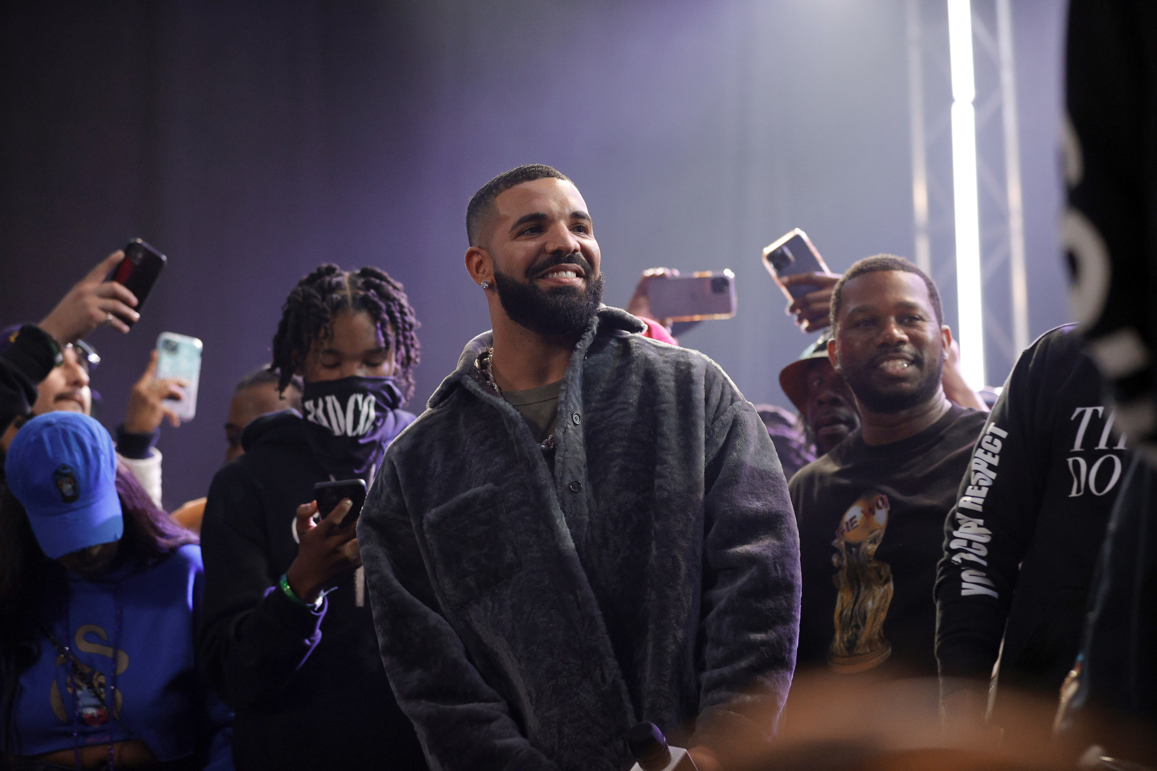 Drake wearing a fur coat smiles among fans holding up phones