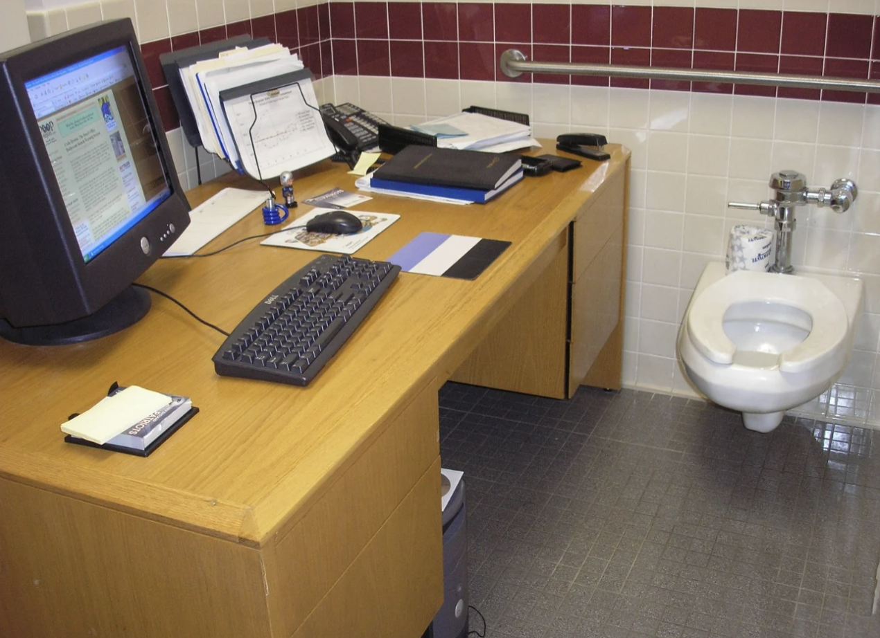 Office desk with computer setup in a bathroom next to a toilet