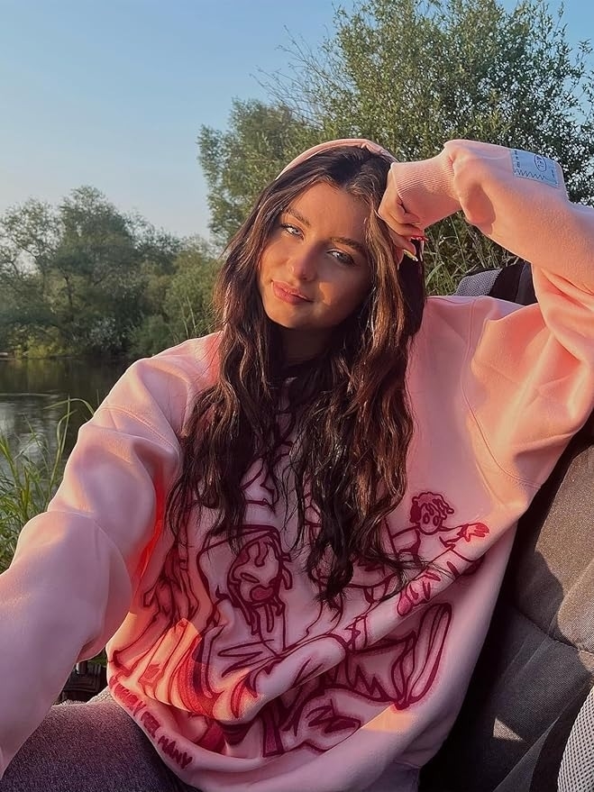 a model in a pink graphic sweatshirt sits outdoors, sunlight highlighting her face