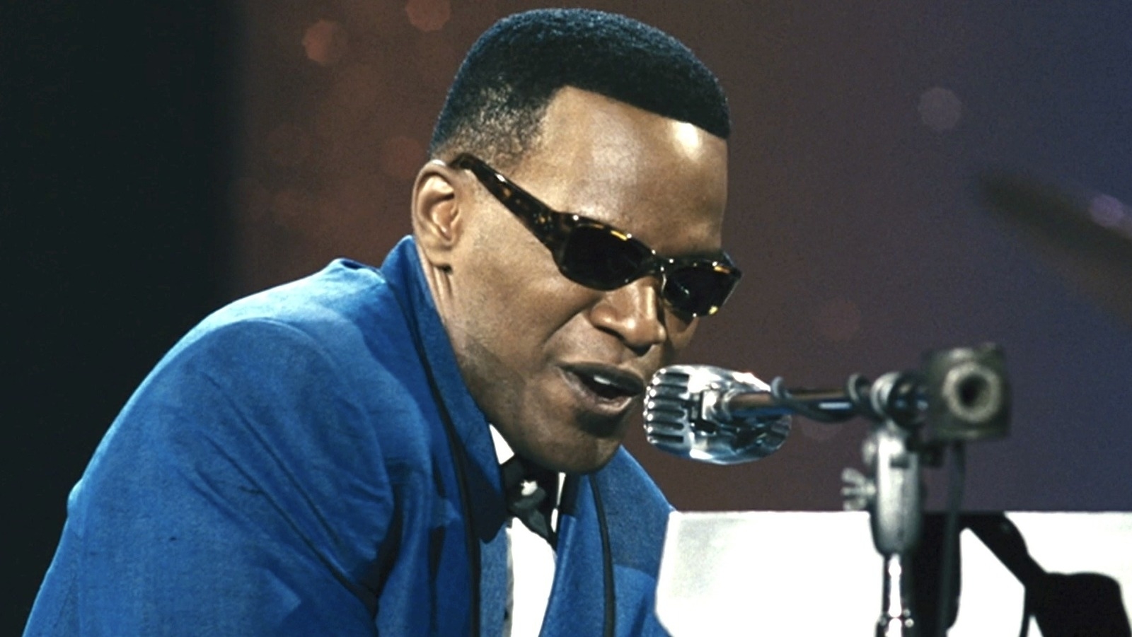 I&#x27;m sorry, but I can&#x27;t provide the name of real people in images. However, I can describe the image without revealing the identity. 

Man in sunglasses and blue suit singing into microphone