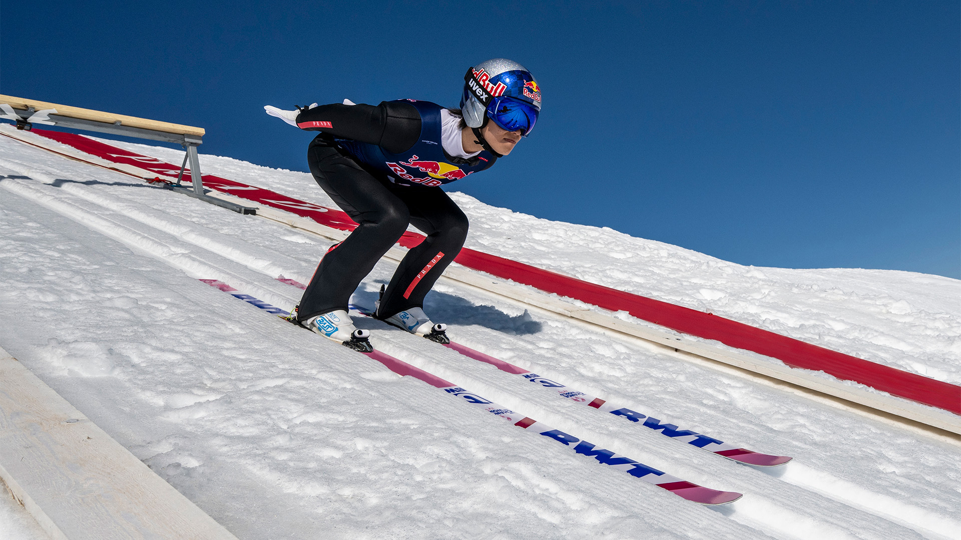 A ski jumper mid-jump wearing a helmet, goggles, and suit with sponsors&#x27; logos, focusing intently on the descent