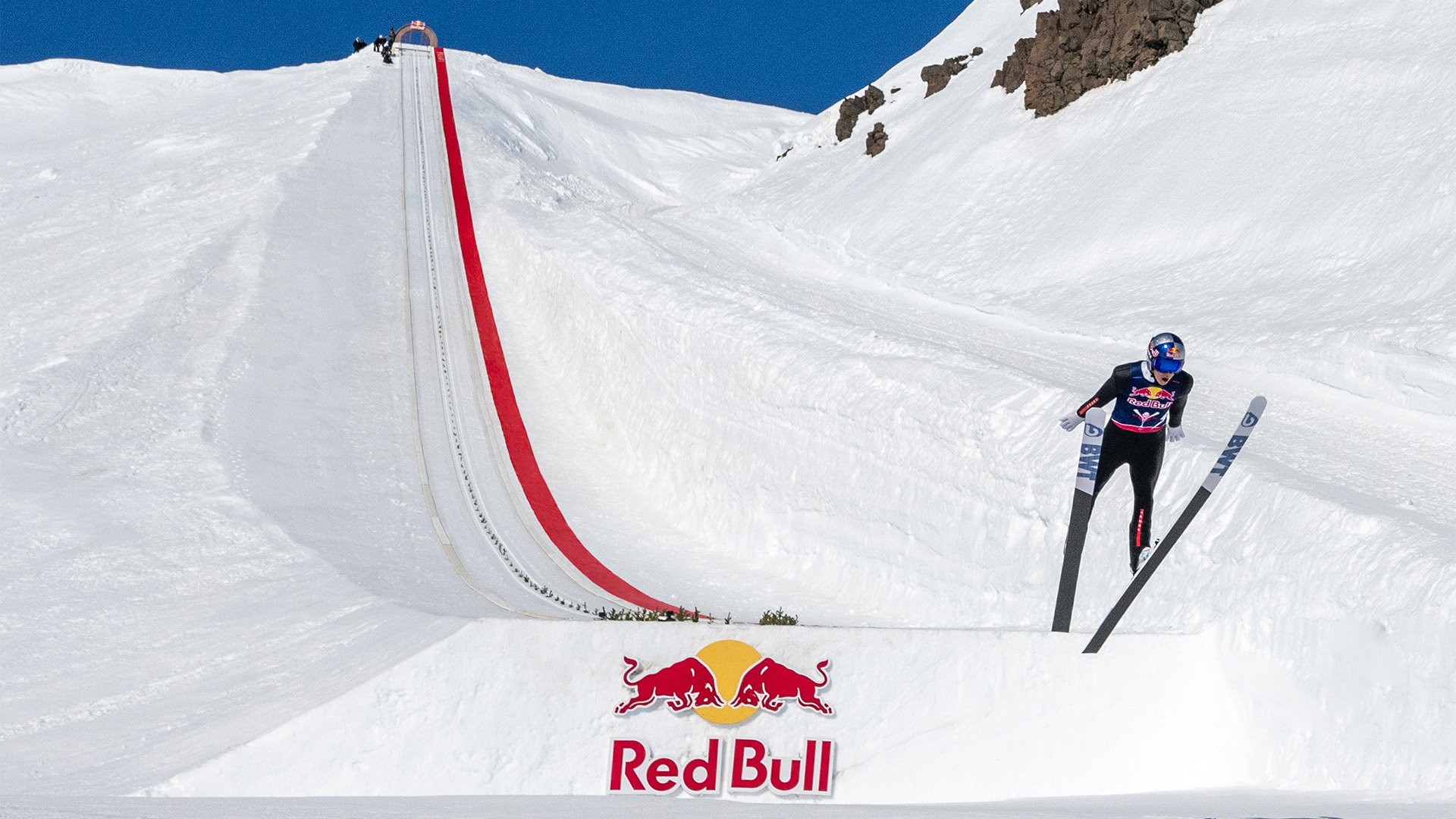Skier mid-jump above a Red Bull banner on a snowy ramp