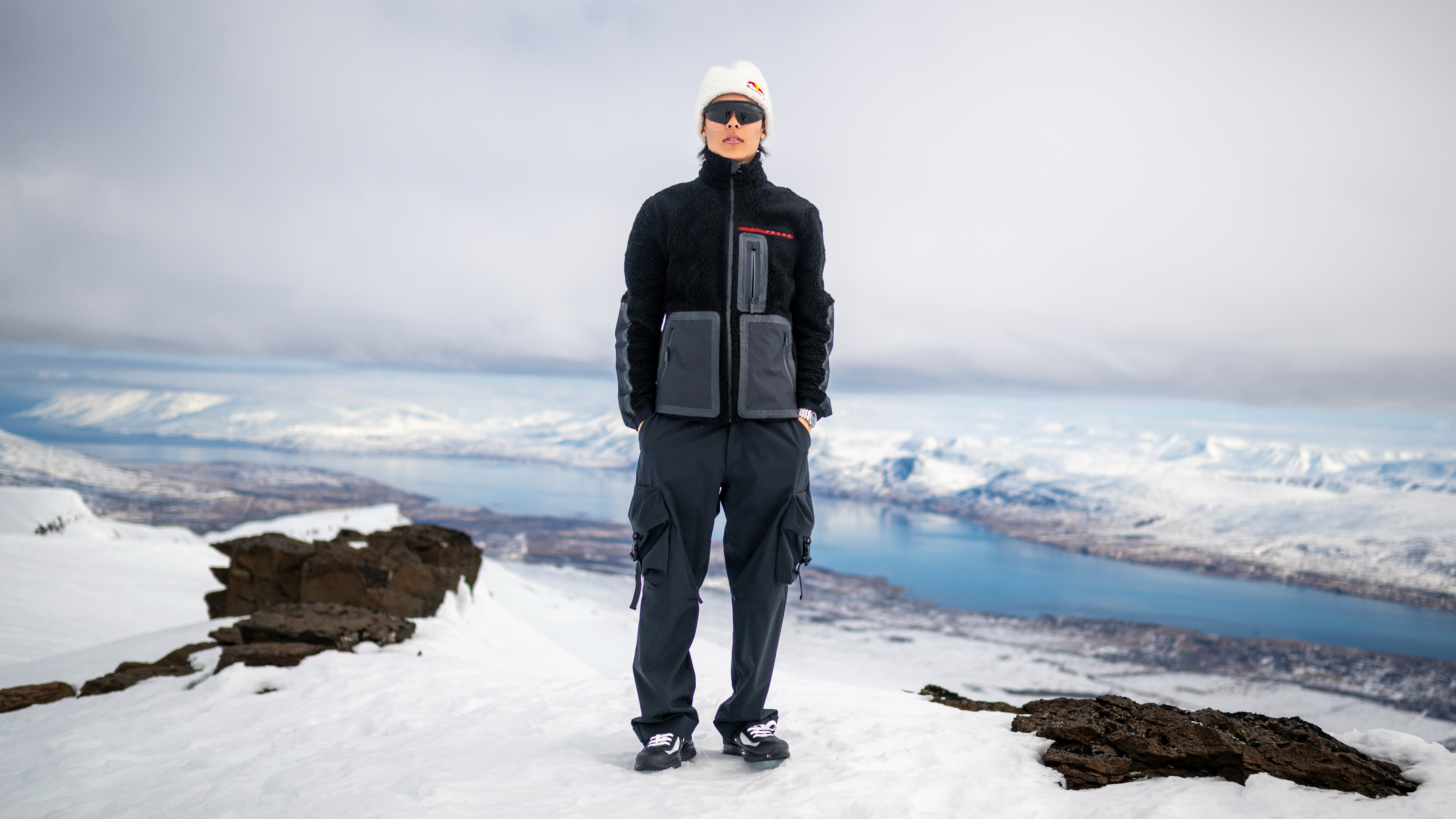 Person stands on snowy overlook in layered outdoor wear with visor cap, against mountainous backdrop