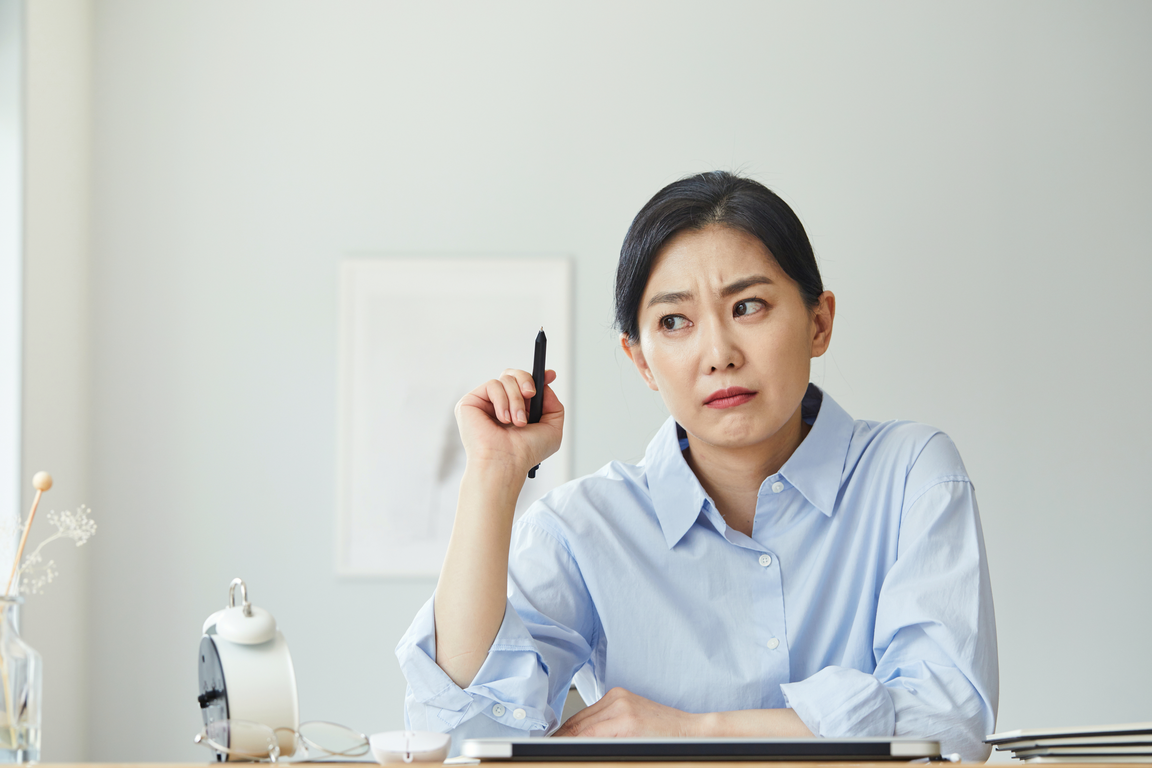 Person seated at a desk, looking puzzled, holding a pen, with papers and a clock in view