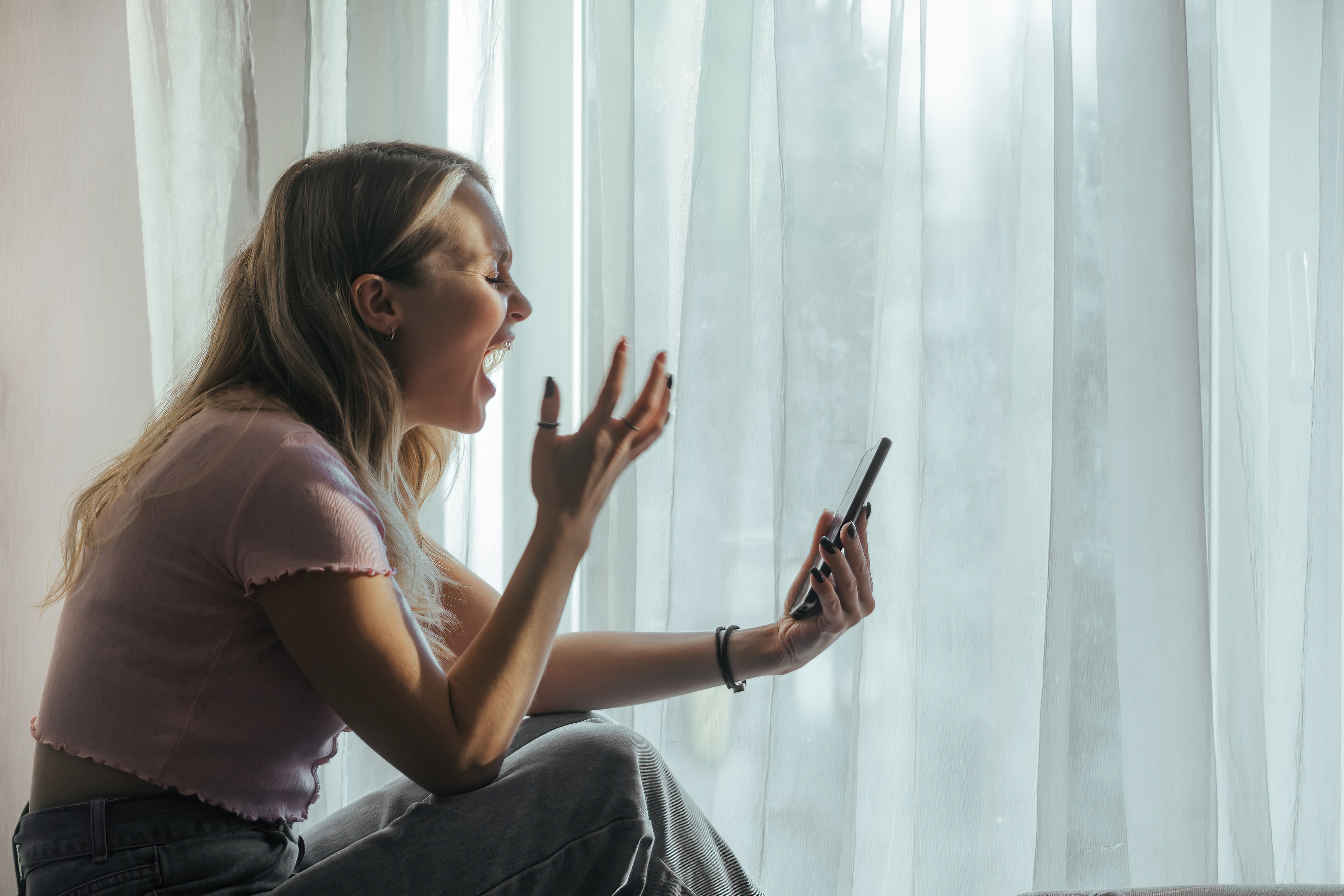 Woman sitting by window, excitedly gesturing at her phone. She&#x27;s in a casual outfit