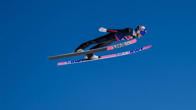 Ski jumper mid-flight against a clear sky, wearing a jumpsuit with sponsor logos
