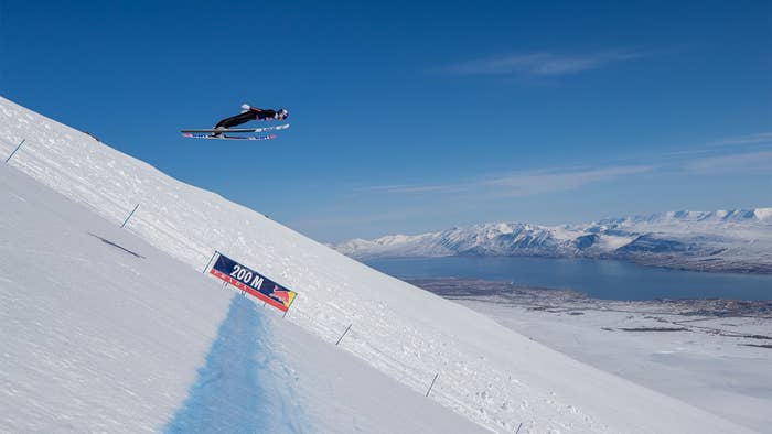 Skier in mid-jump over a snowy slope, with a clear sky and snow-covered mountains in the background