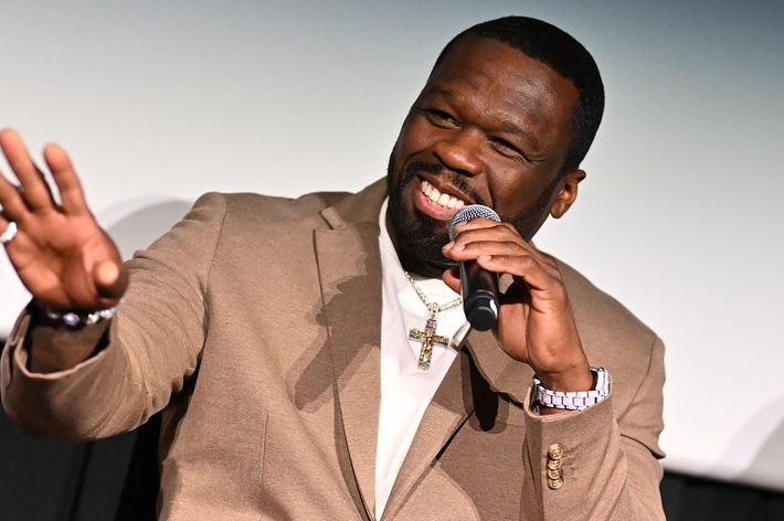 50 Cent in beige suit smiling and gesturing while speaking into a microphone