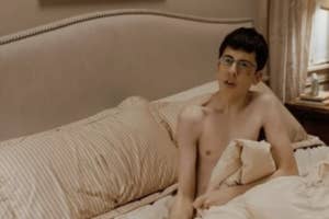 Man with glasses sitting in bed, topless, looking surprised or caught off guard