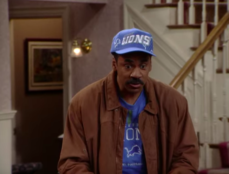 Man wearing a baseball cap and jacket stands in a room with a staircase in the background