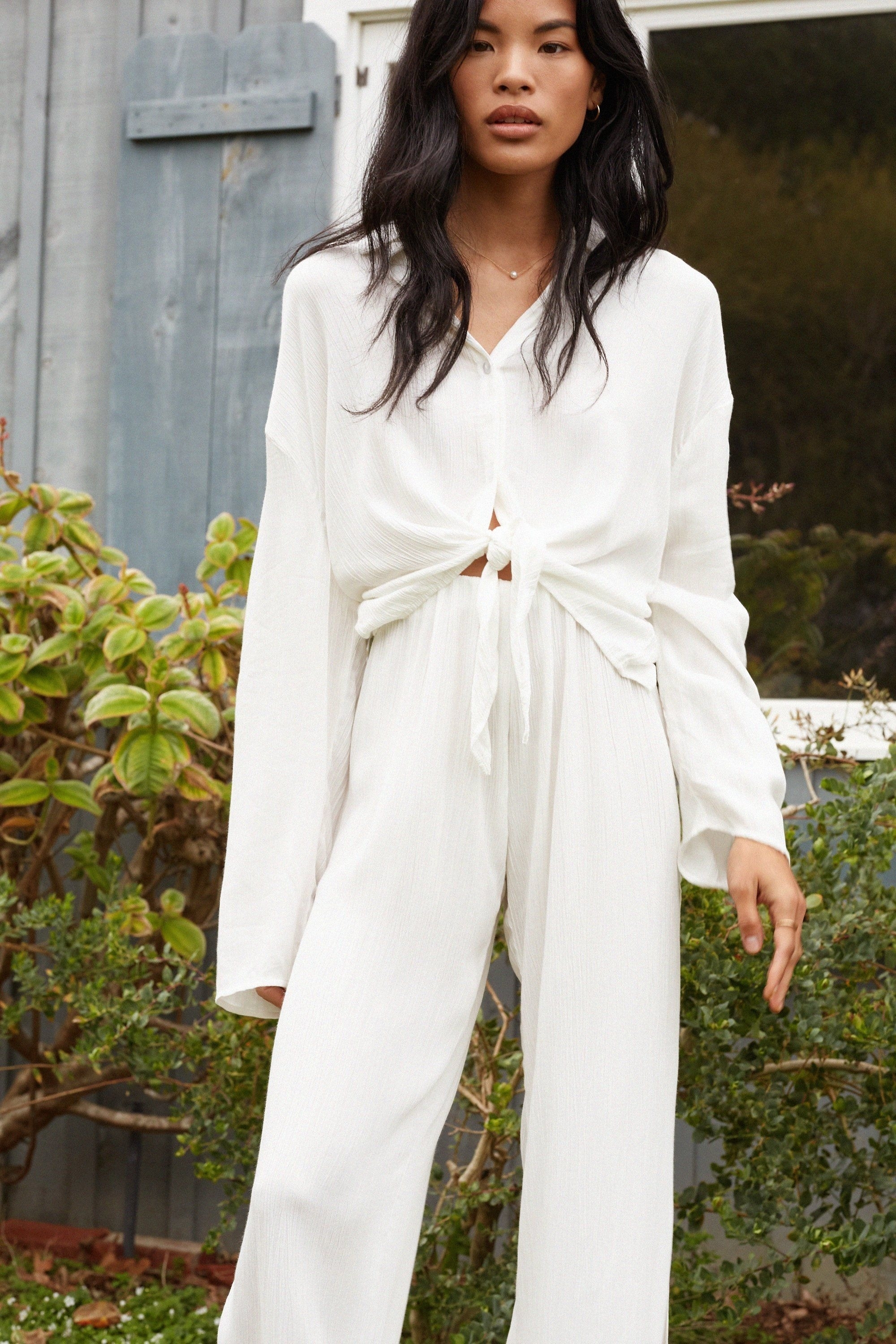 Woman in a casual white tie-front jumpsuit with long sleeves, standing in a garden for a fashion ad