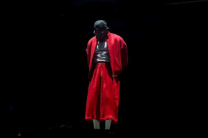 Music artist on stage wearing a shiny top and red robe, immersed in a dramatic spotlight