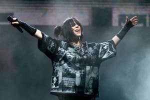 Billie Eilish performing on stage, wearing a graphic oversized shirt and gloves, with open arms and a joyous expression