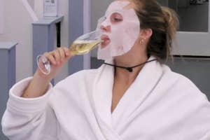 Woman in a robe with a facial mask sipping a drink, embodying self-care relaxation