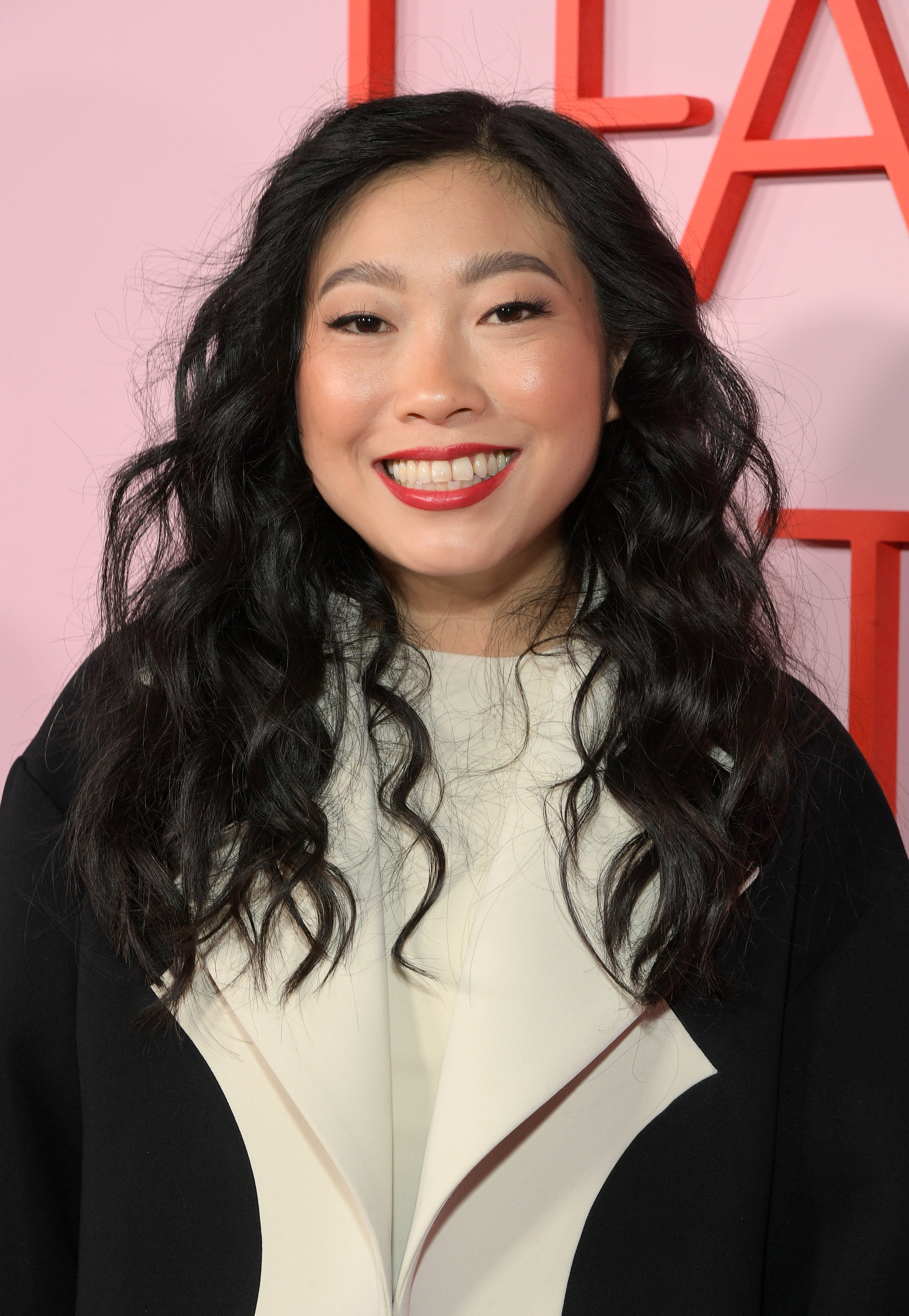 Awkwafina smiling at event, wearing a white top with black jacket, subtle makeup and wavy hair