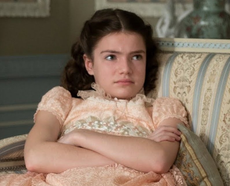 Joven actriz en vestido vintage sentada en sofá, con expresión pensativa y cruzando los brazos