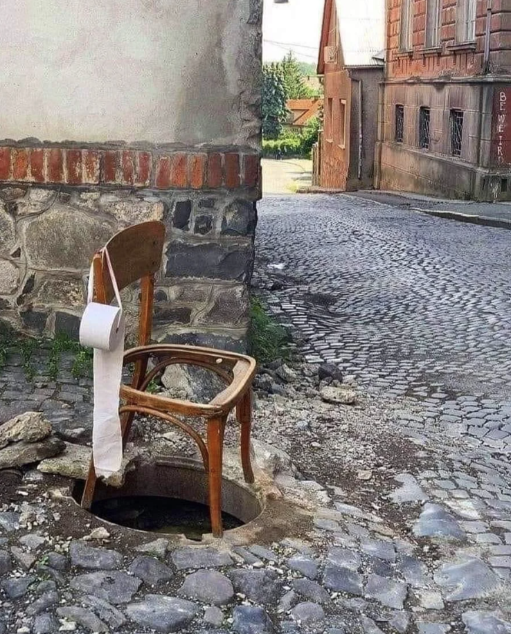 A makeshift toilet comprising a chair with a hole placed over an open manhole, with toilet paper attached