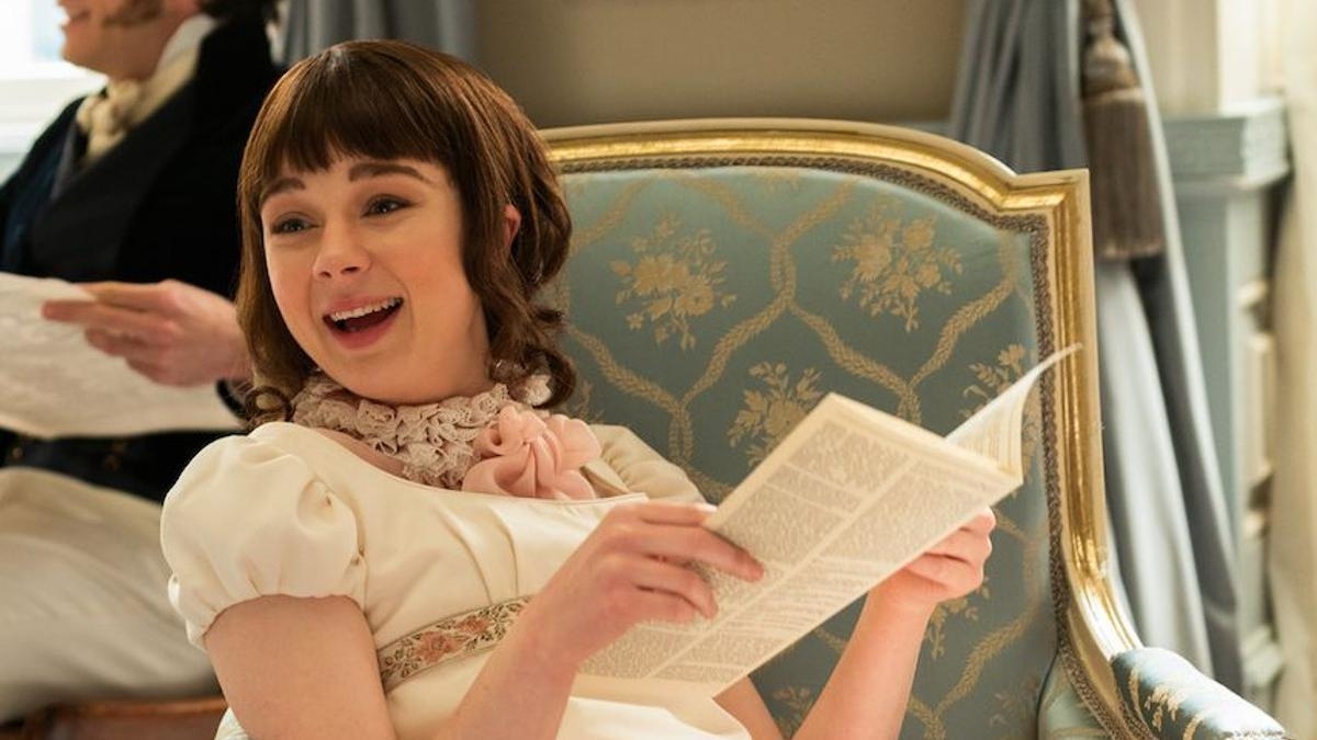 Personaje Eloise Bridgerton de la serie de televisión sonríe sentado en una silla elegante, sostiene un periódico