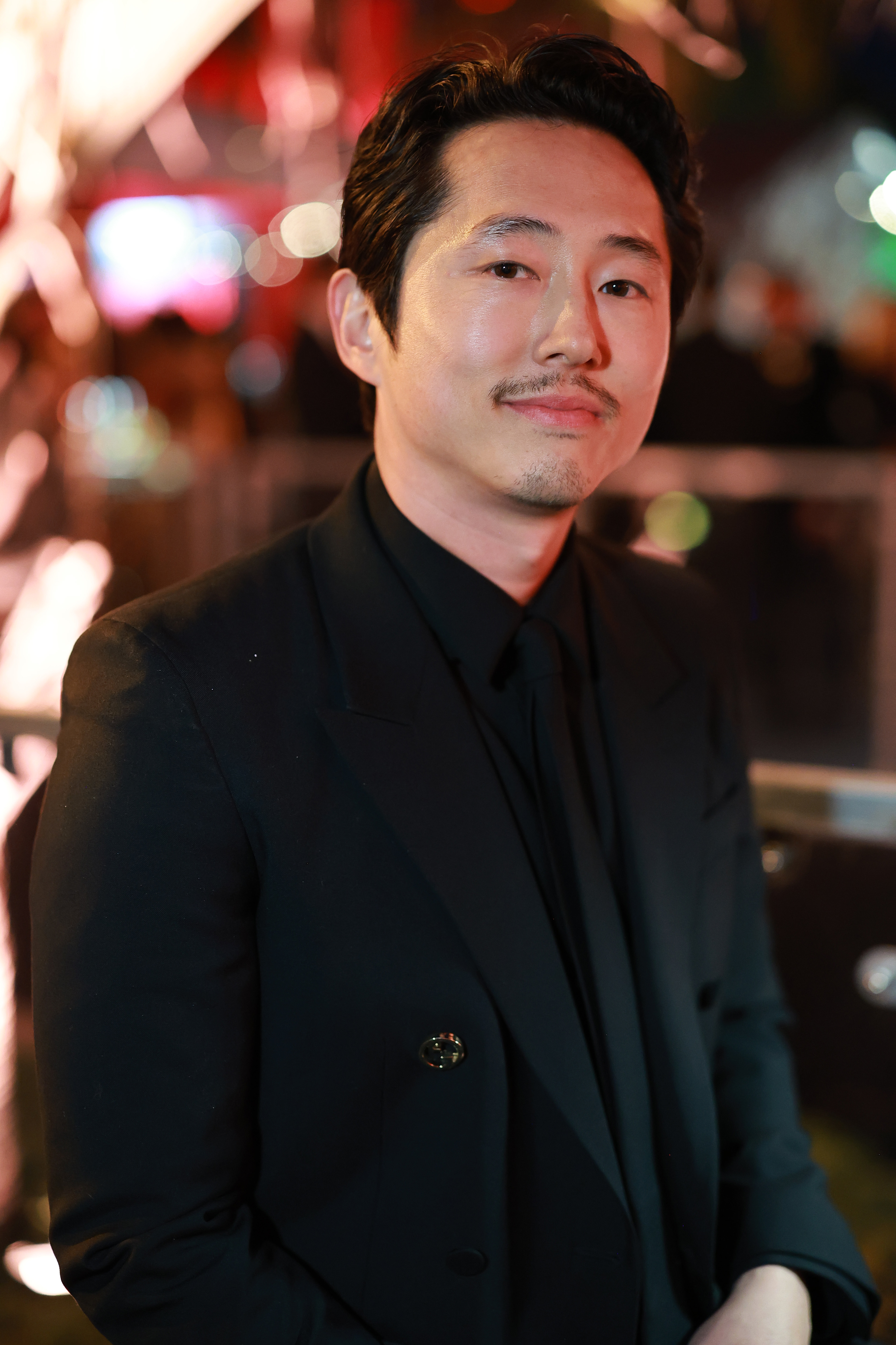 Steven Yeun in a formal black suit smiling at a celebrity event