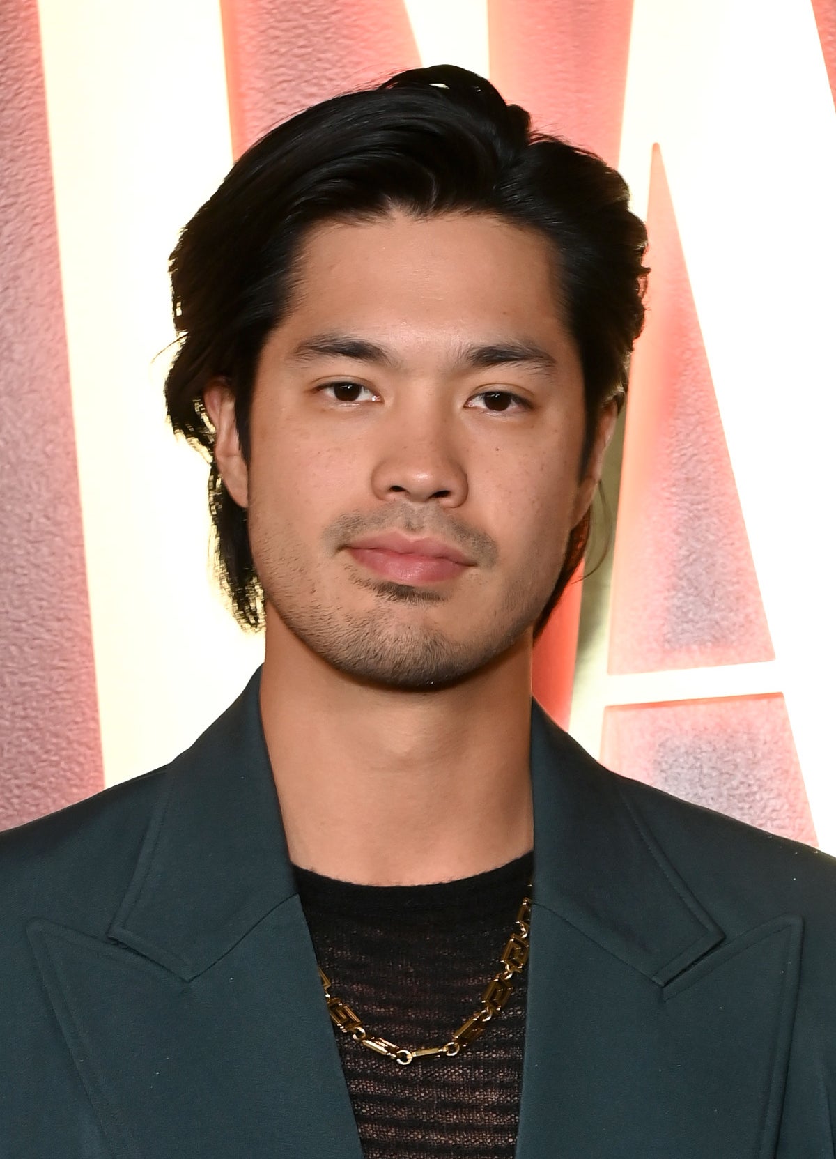 Ross Butler posing at event, wearing a blazer over a mesh top with layered necklaces