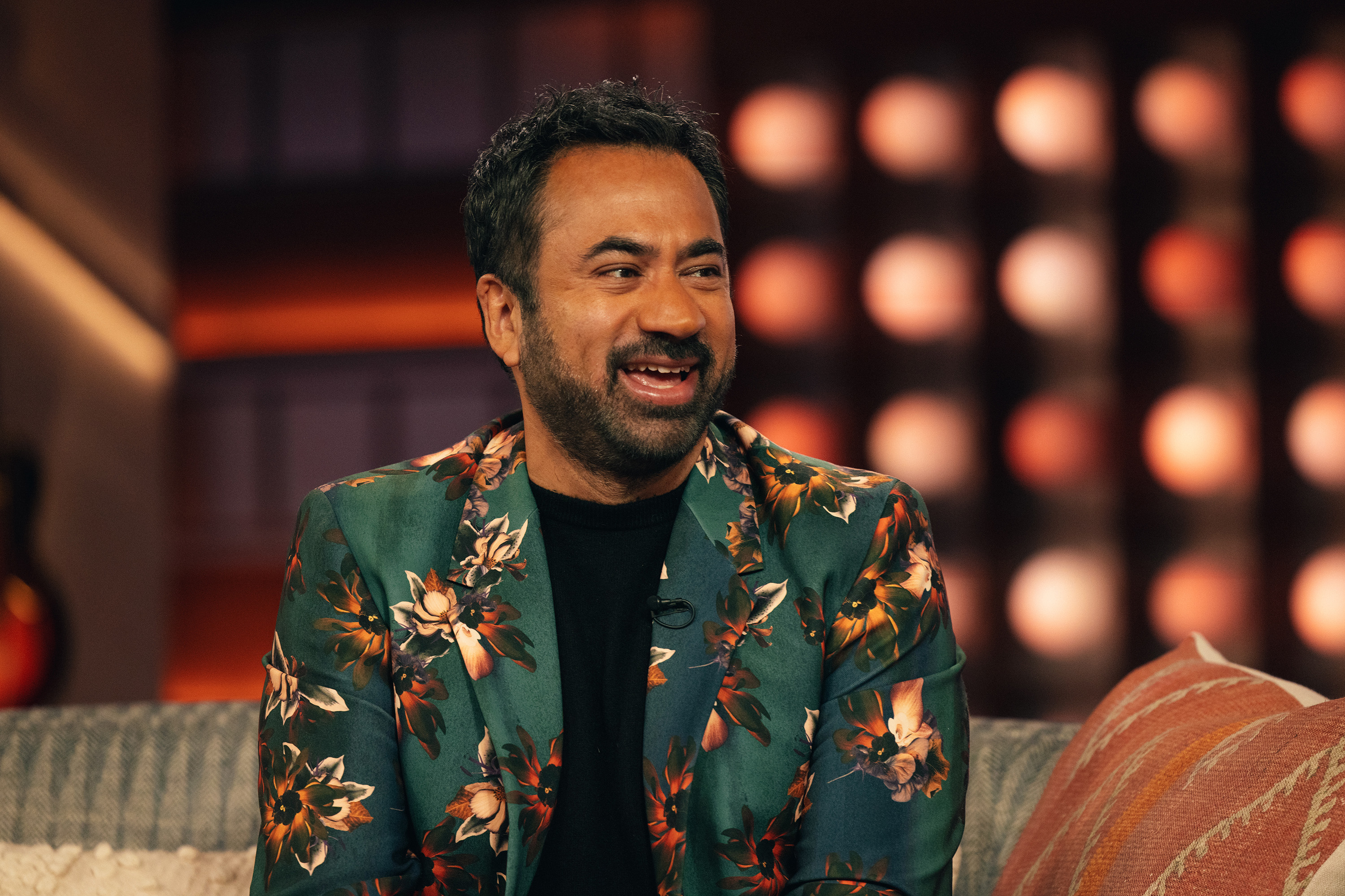 Kal Penn in a floral jacket sitting with a smile on a talk show set