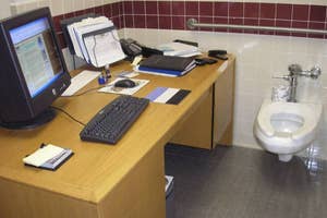 Office desk with computer setup next to a toilet, blending work and personal space uniquely