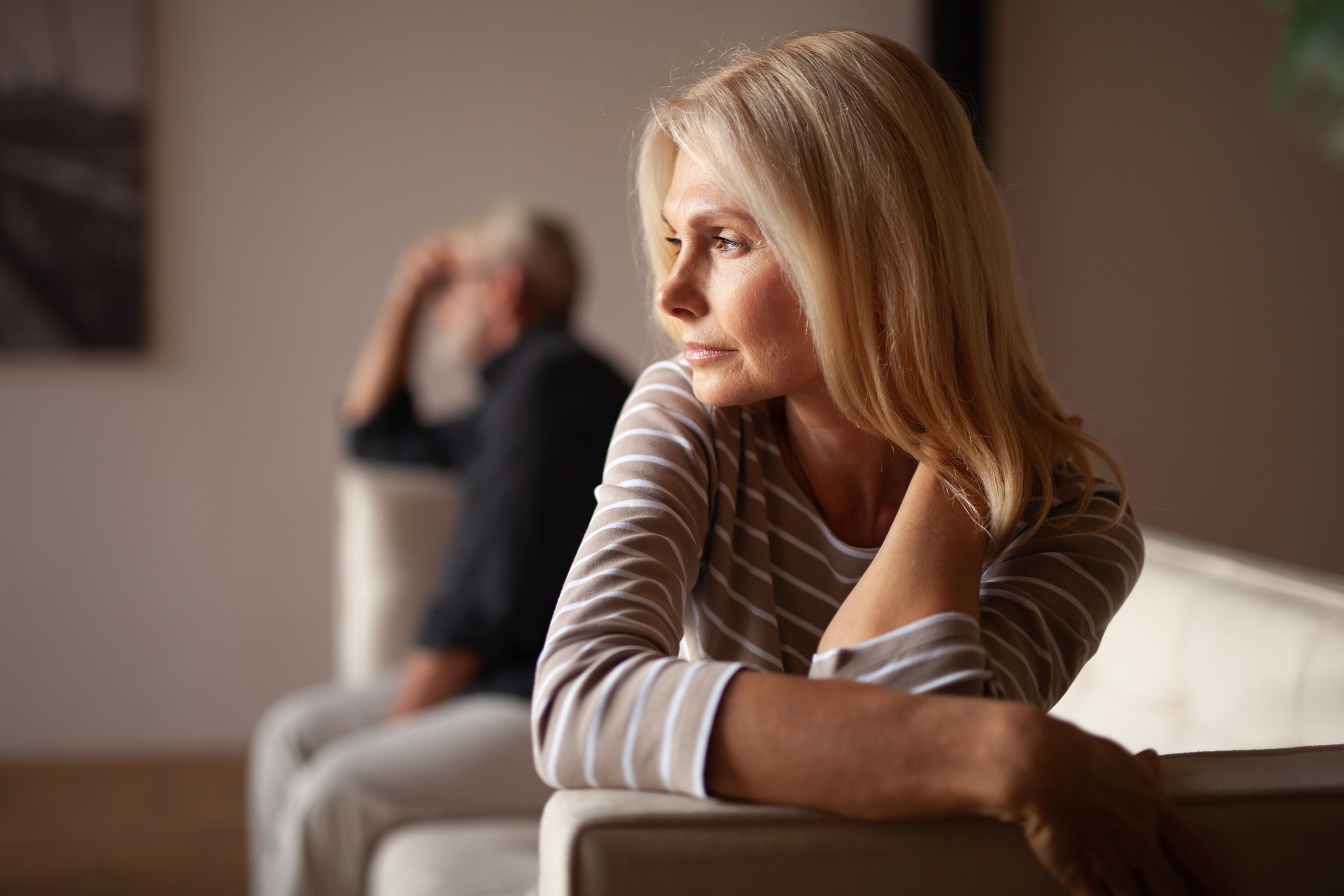 A woman looking thoughtful in the foreground, with a man sitting in the background out of focus, both appearing contemplative