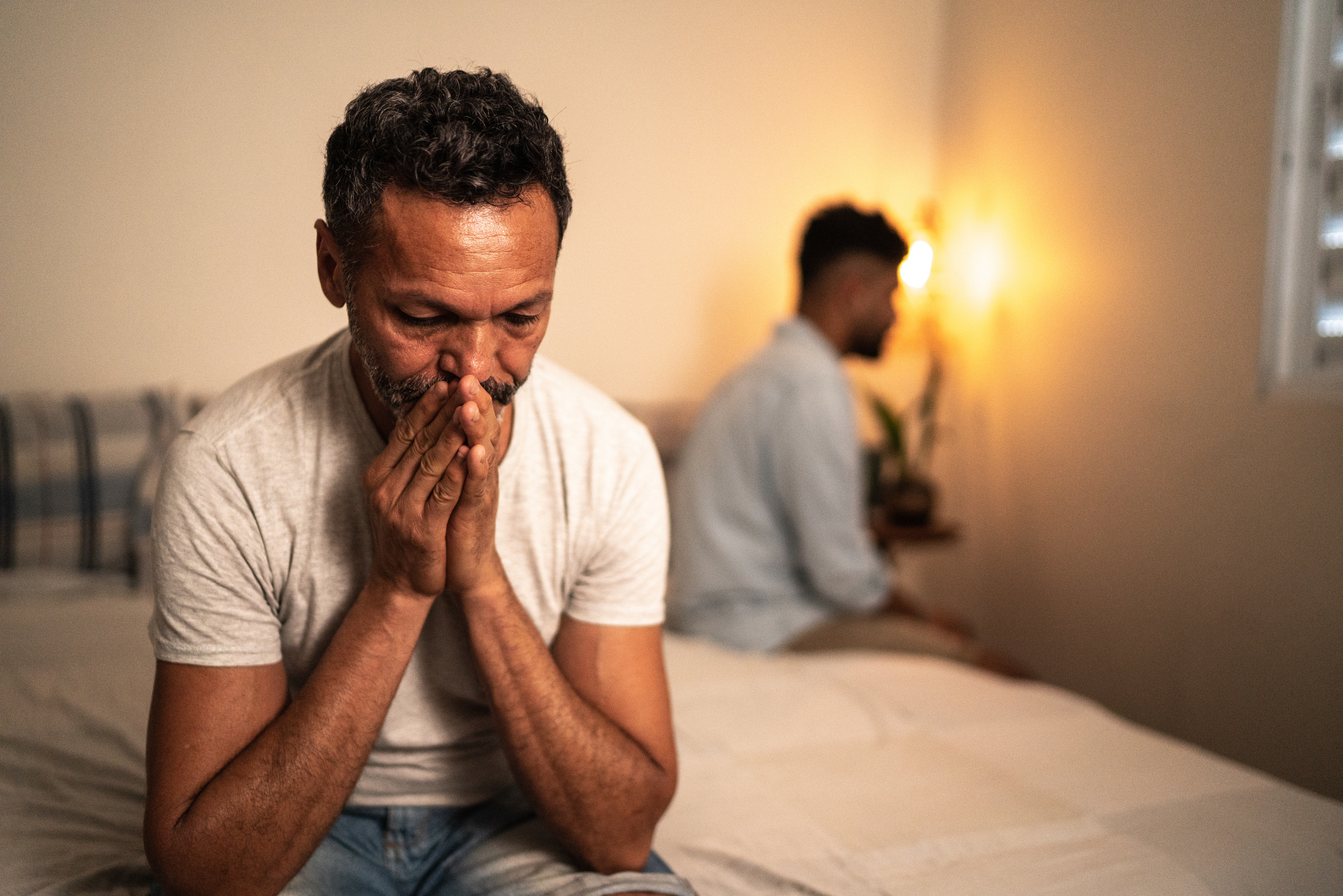 Two men sitting separately on a bed, one with hands clasped, suggesting a disagreement or tension