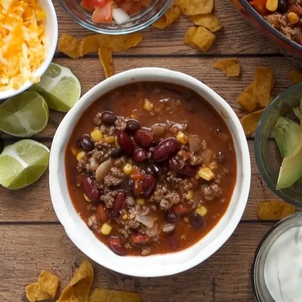 Bowl of chili with beans, corn, and ground meat, surrounded by toppings and chips