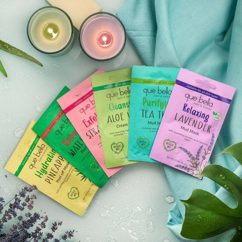 Que Bella face mask packets arranged with candles and leaves, suggesting a spa-like setting for a skincare article
