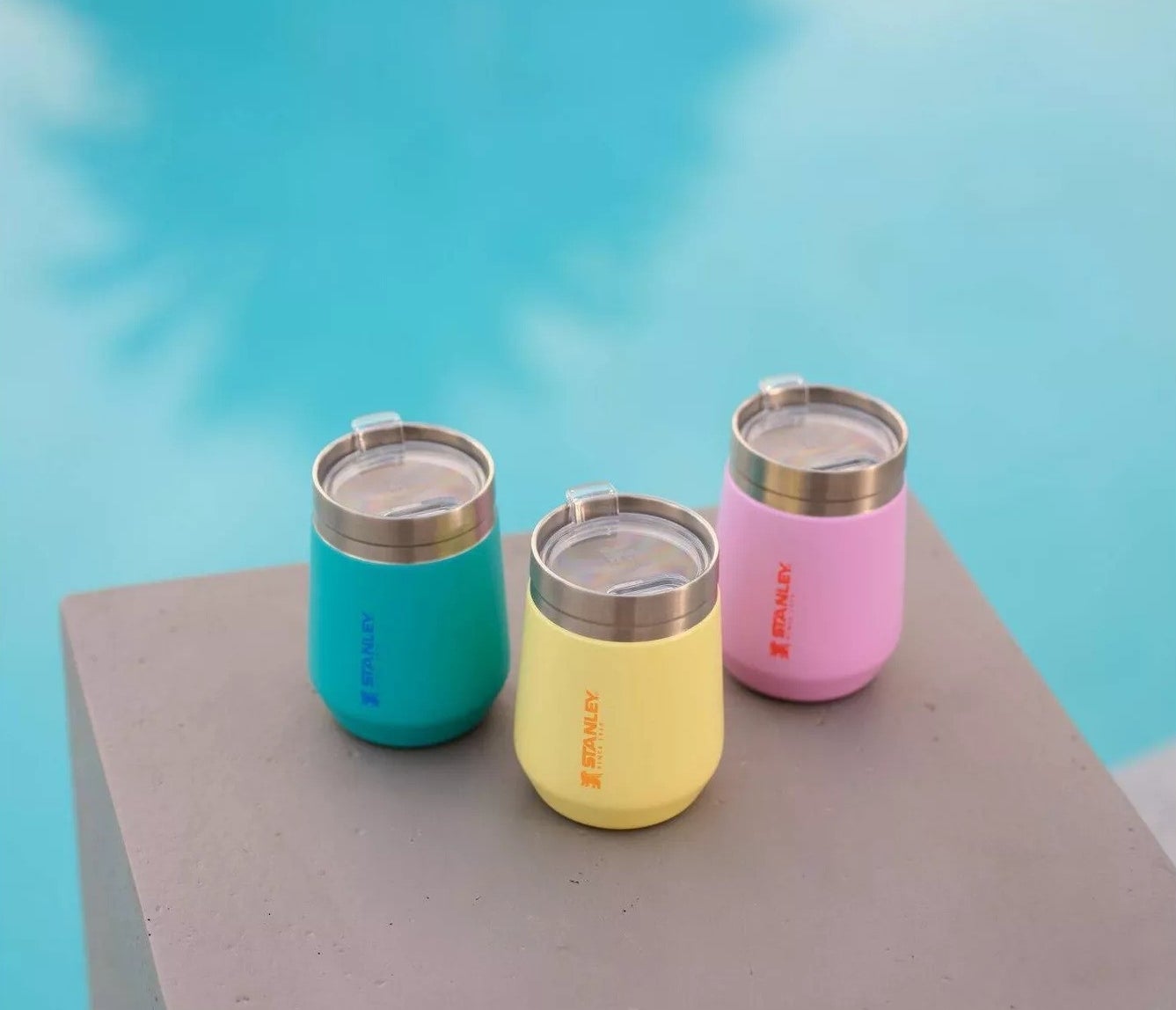 Three insulated tumblers beside a pool, in teal, yellow, and pink, with lids. Ideal for outdoor use